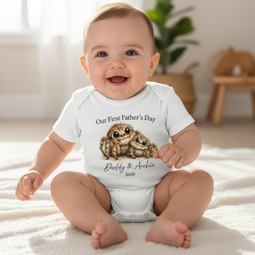 Baby wearing a white bodysuit with two huntsman spiders and Our First Father's Day text