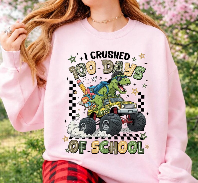 A person wearing a light pink sweatshirt with a colorful graphic that reads 'I CRUSHED 100 DAYS OF SCHOOL' featuring a green dinosaur driving a yellow monster truck. The background is a blurred outdoor garden setting.