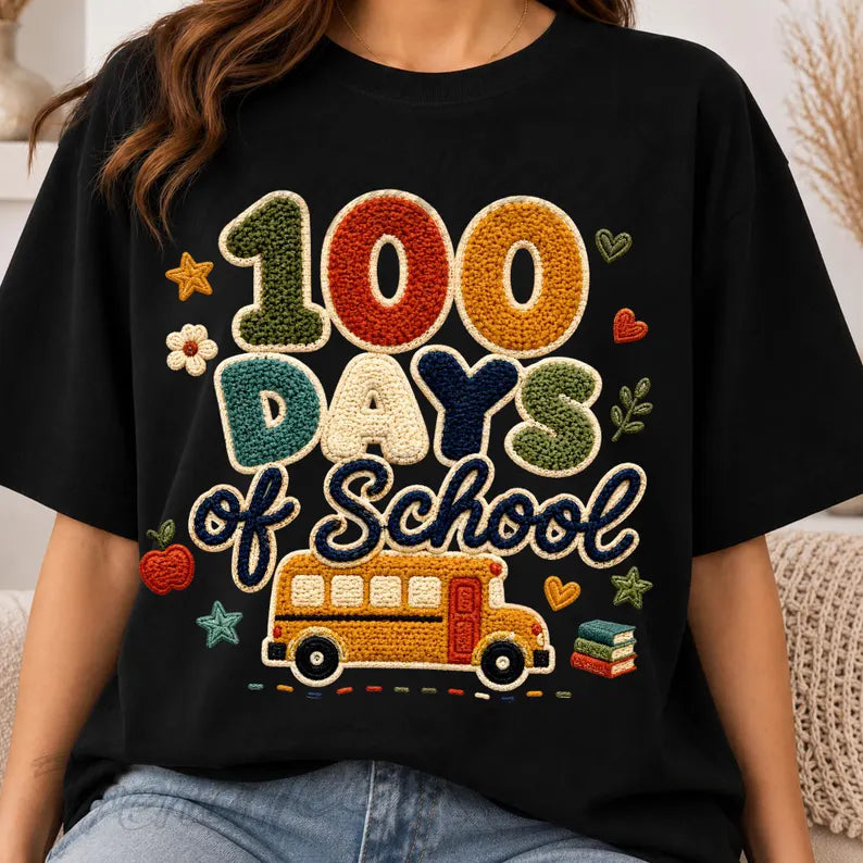 A person wearing a black short-sleeve T-shirt with a large colorful embroidered-style graphic reading "100 Days of School" above a yellow school bus, shown in a real indoor setting.