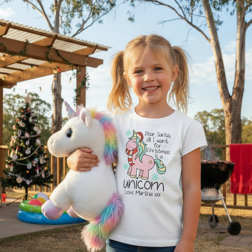 A white t-shirt with a graphic print of a unicorn, Santa Claus, and text that reads 'Dear Santa, All I want for Christmas is a UNICORN' along with 'Love Martha'.