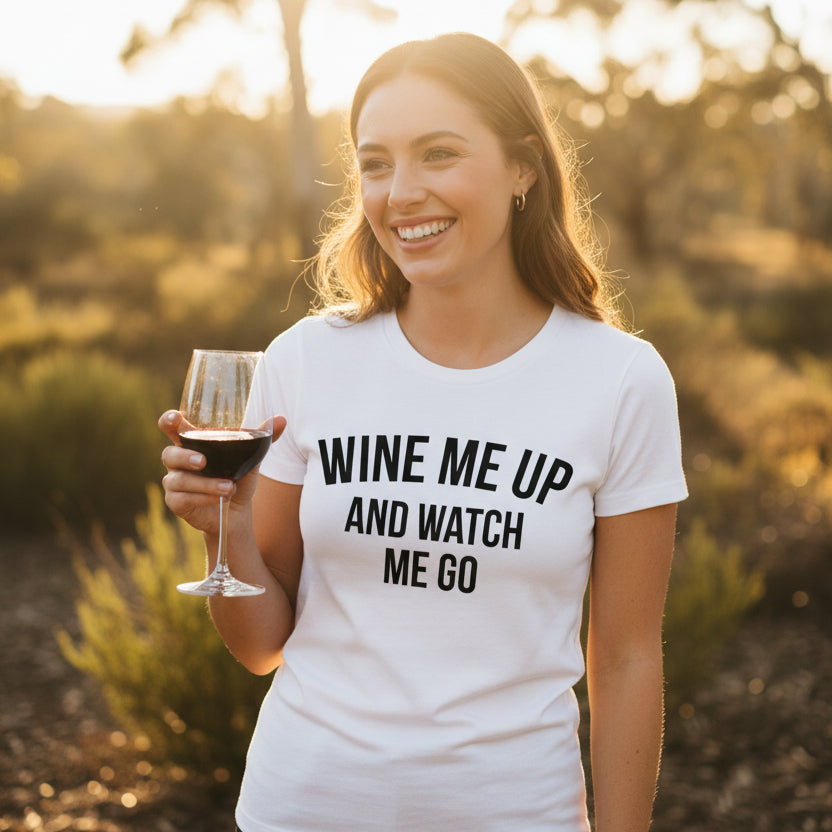 Smiling woman holding a glass of red wine outdoors wearing a white Wine Me Up And Watch Me Go t-shirt with bold black lettering