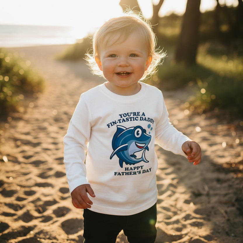 Toddler at the beach wearing a white long-sleeve t-shirt with a cartoon shark reading You're A Fin-Tastic Daddy, Happy Father's Day.