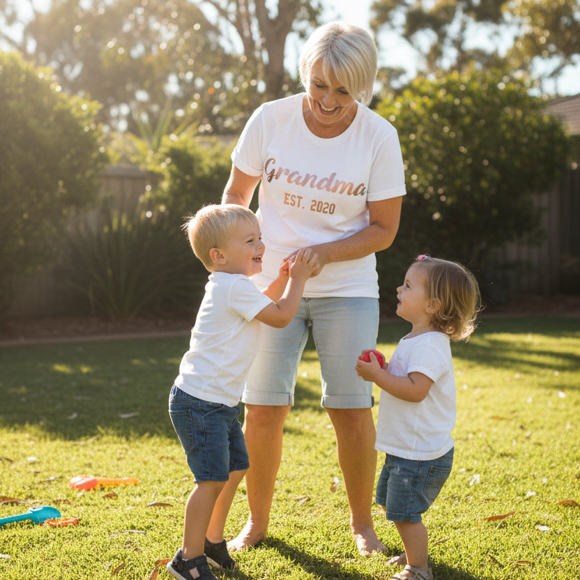 White womens t-shirt with personalised Grandma Est. 2020 design in rose gold script and block lettering laid flat with peonies