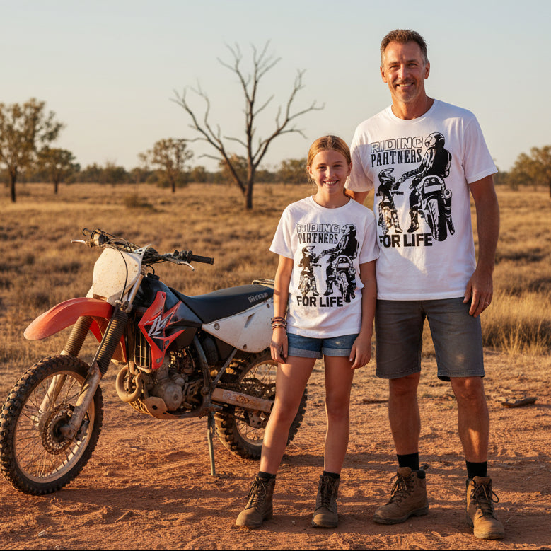 Two white t-shirts laid flat showing matching Riding Partners For Life design with dad and daughter on motorbikes fist bumping in bold black silhouette