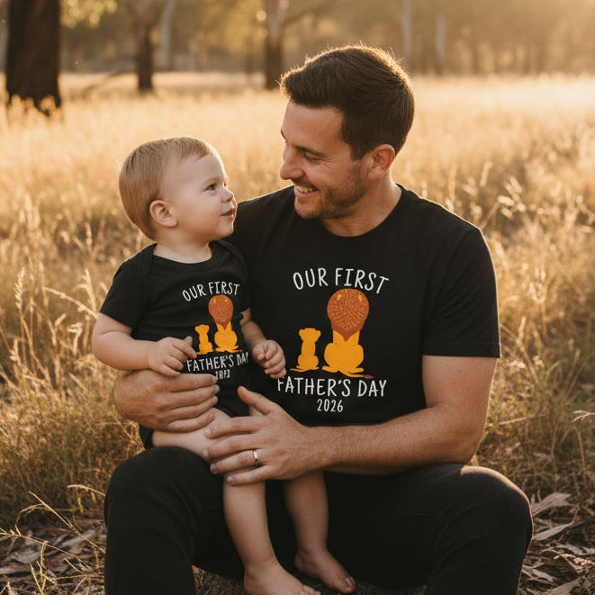 Dad and baby sitting in a field wearing matching black tops with a lion and cub Our First Father's Day design and the year.