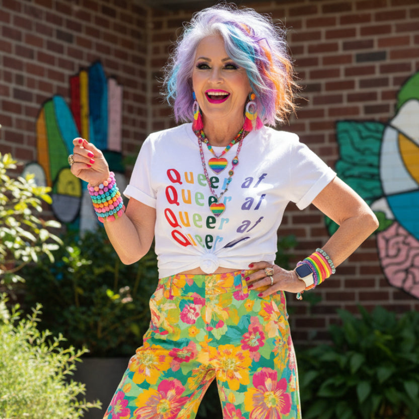 White t-shirt with 'Queer at' text in rainbow colors on a light background.