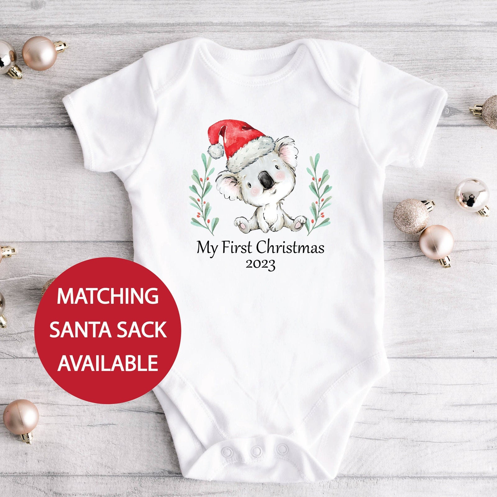 White baby onesie with a Christmas design of a koala wearing a Santa hat, on a light wooden surface with Christmas ornaments.