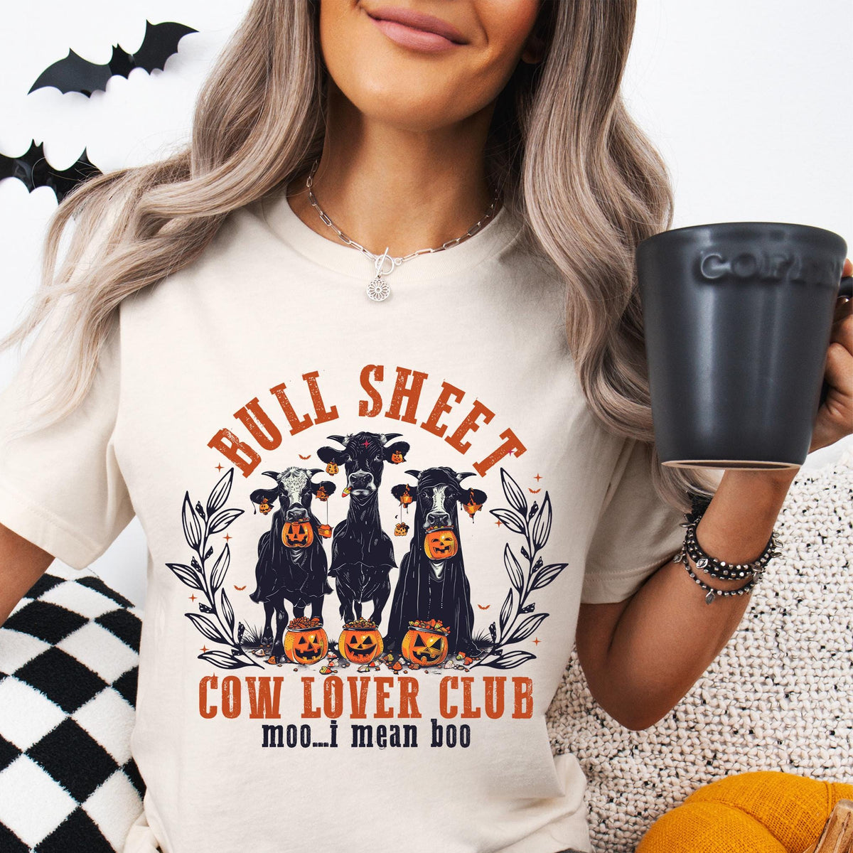 Person wearing a &#39;Bull Sheet Cow Lover Club&#39; t-shirt with Halloween design, holding a mug.