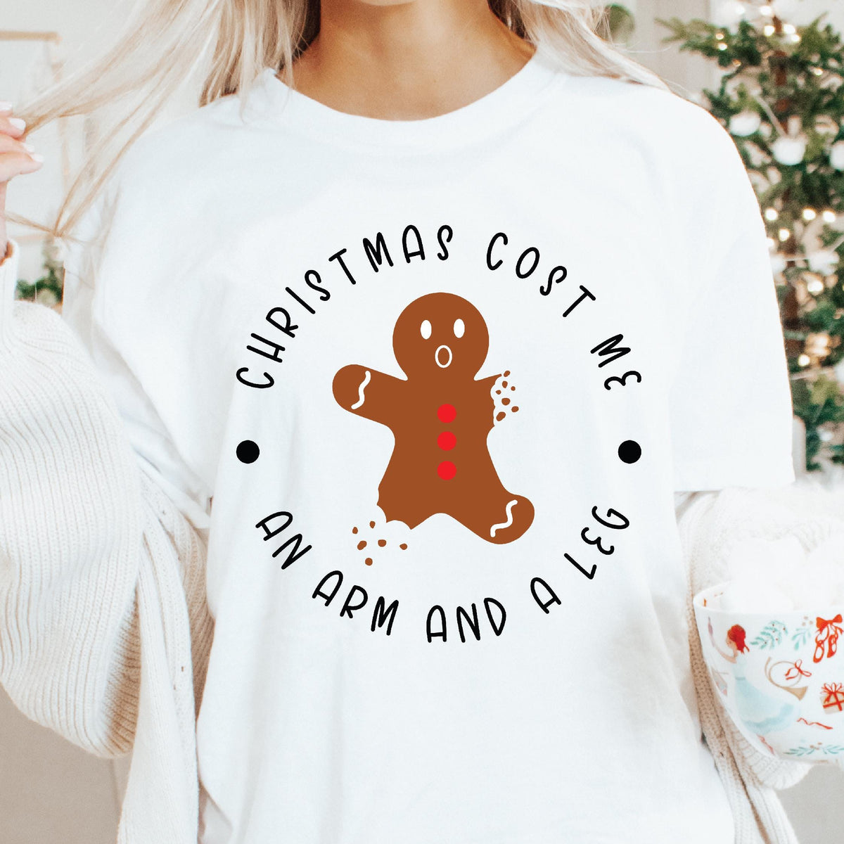 A person holding a mug and wearing a white sweatshirt with a Christmas-themed graphic that reads 'Christmas Cost Me an Arm and a Leg' and depicts a gingerbread man. The background indicates a cozy, indoor Christmas setting.