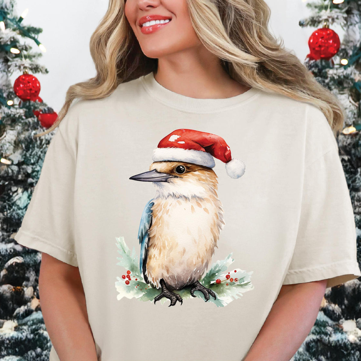 A person wearing a beige t-shirt with a printed image of a kookaburra wearing a Santa hat, surrounded by Christmas decorations.