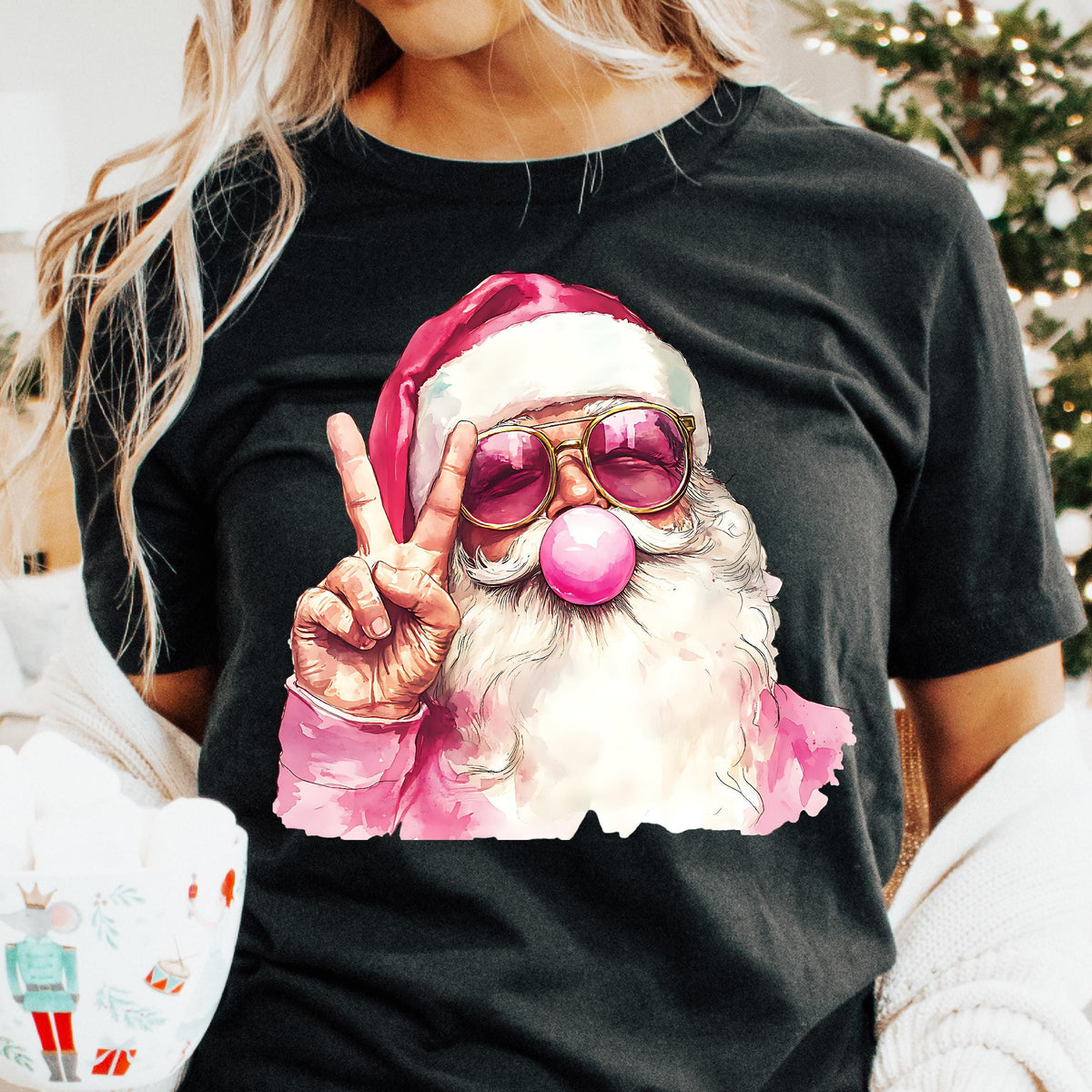 A person wearing a black t-shirt with a colorful illustration of a smiling Santa Claus making a peace sign, surrounded by a cozy indoor setting with Christmas decorations.