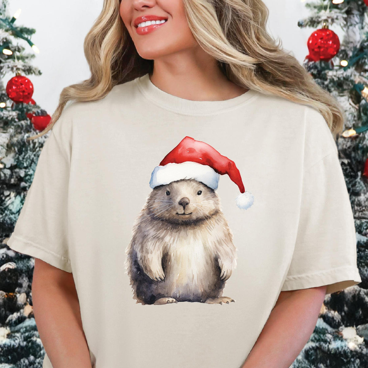 A person wearing a beige t-shirt with a printed image of a wombat wearing a Santa hat, surrounded by Christmas decorations.
