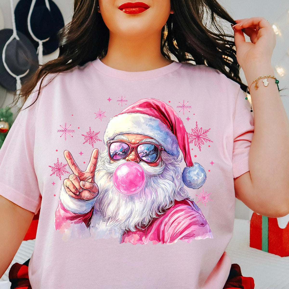 A person wearing a pink t-shirt with a printed design of Santa Claus in sunglasses and a bubble, indicating the product is available in various colors.