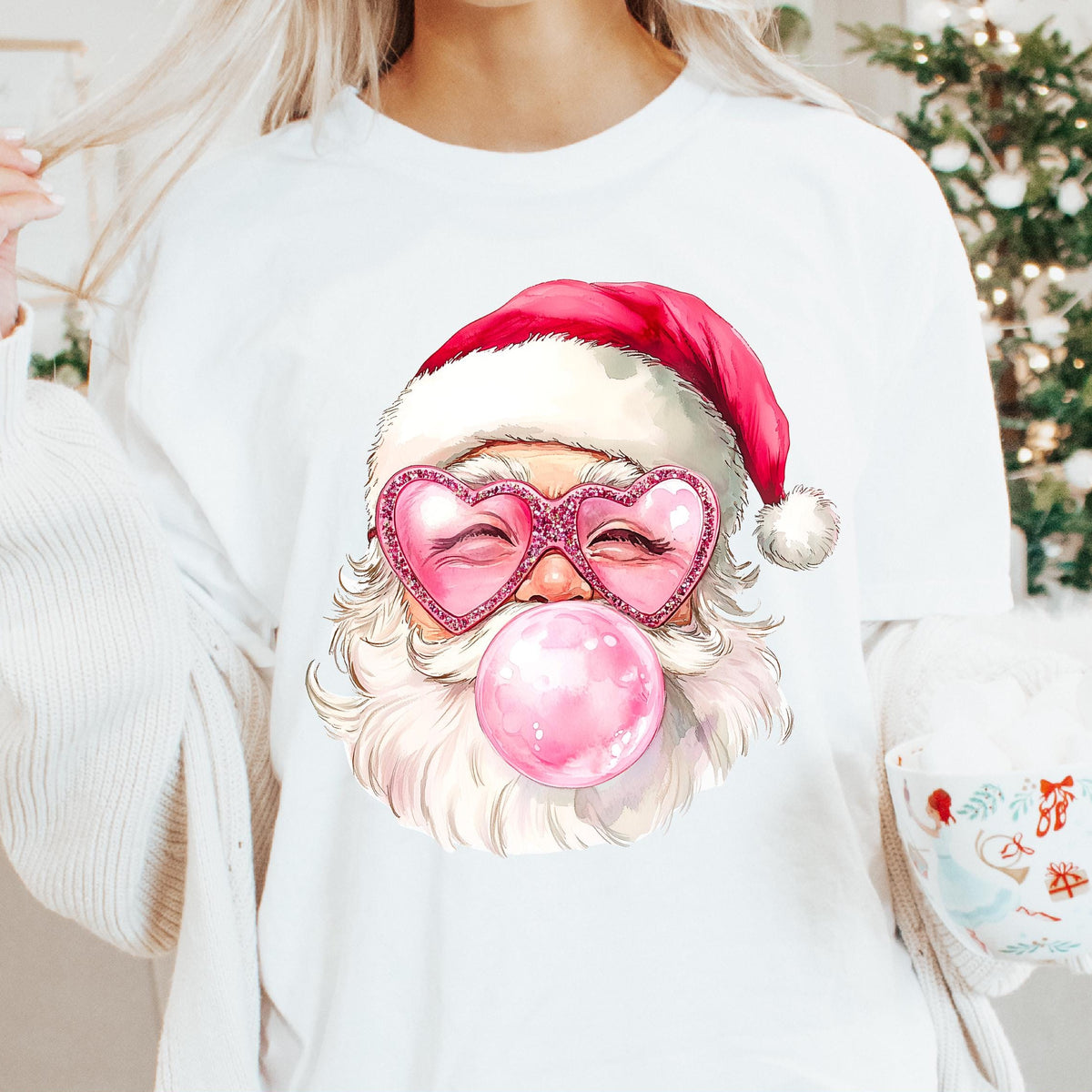 A woman wearing a white sweatshirt with a colorful illustration of Santa Claus wearing a pink Santa hat and a pink bubble gum, holding a mug, and surrounded by Christmas decorations.