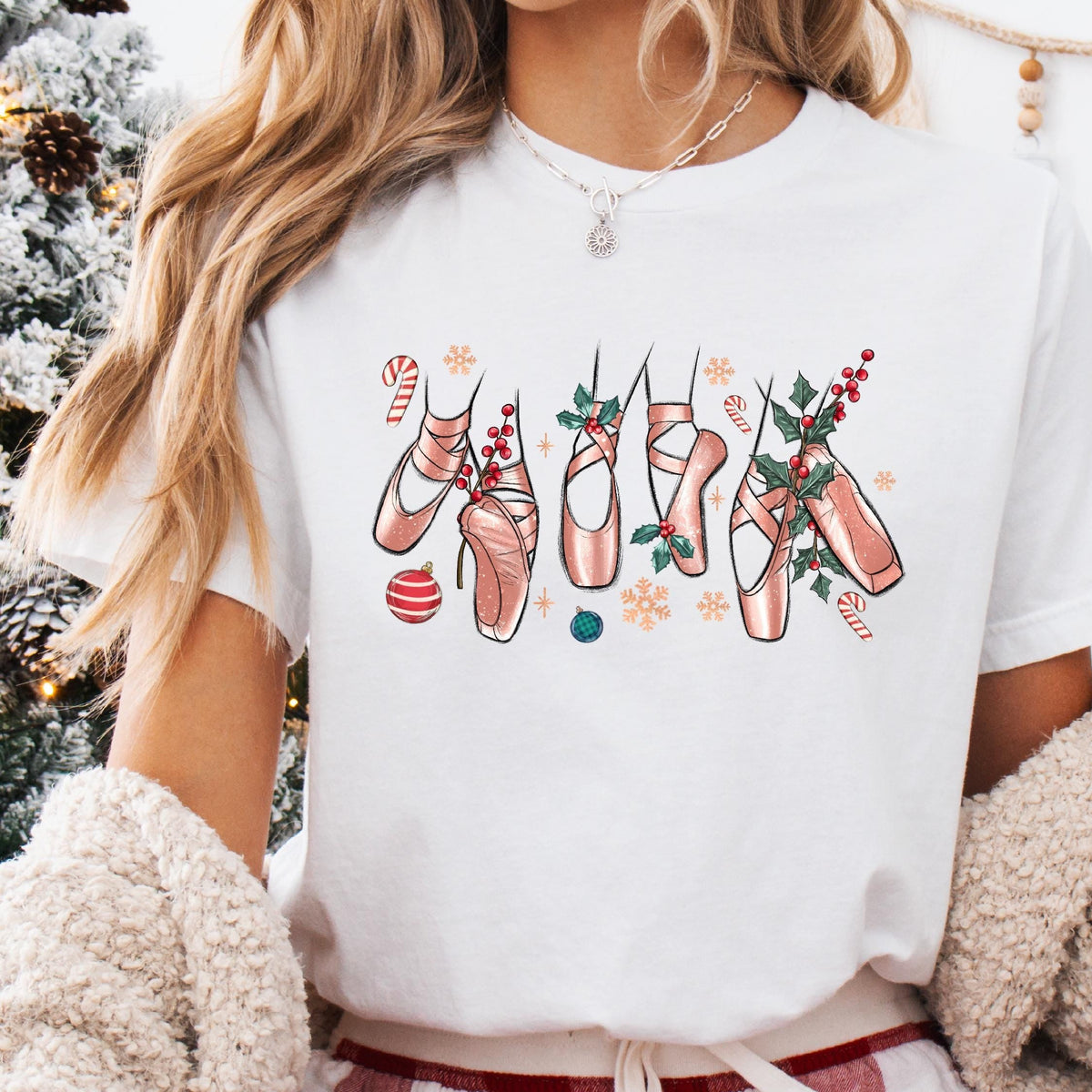 A person wearing a white t-shirt with a Christmas-themed ballet slipper design, sitting on a cozy surface with a festive background.