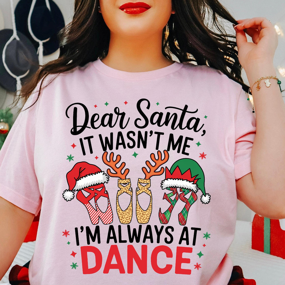 A person wearing a pink t-shirt with 'Dear Santa It Wasn't Me I'm Always At Dance' text and Christmas themed decorations such as Santa hats and reindeer. The shirt is presented in a casual, lying flat on a surface with a blurred background featuring Christmas decorations.