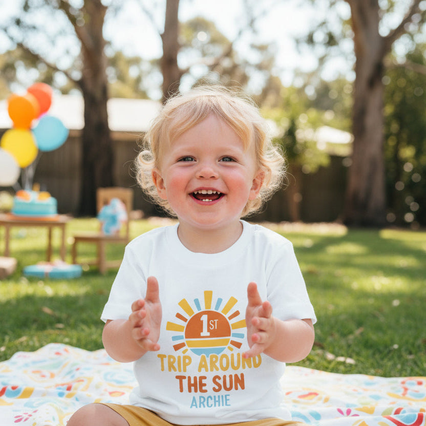 Two white shirts with 'Daddy', 'Mama', and '1st Trip Around the Sun Archie' text on a wooden surface.