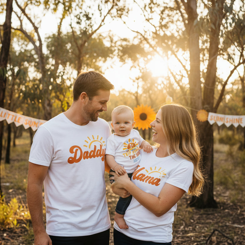Two white shirts with 'Daddy', 'Mama', and '1st Trip Around the Sun Archie' text on a wooden surface.
