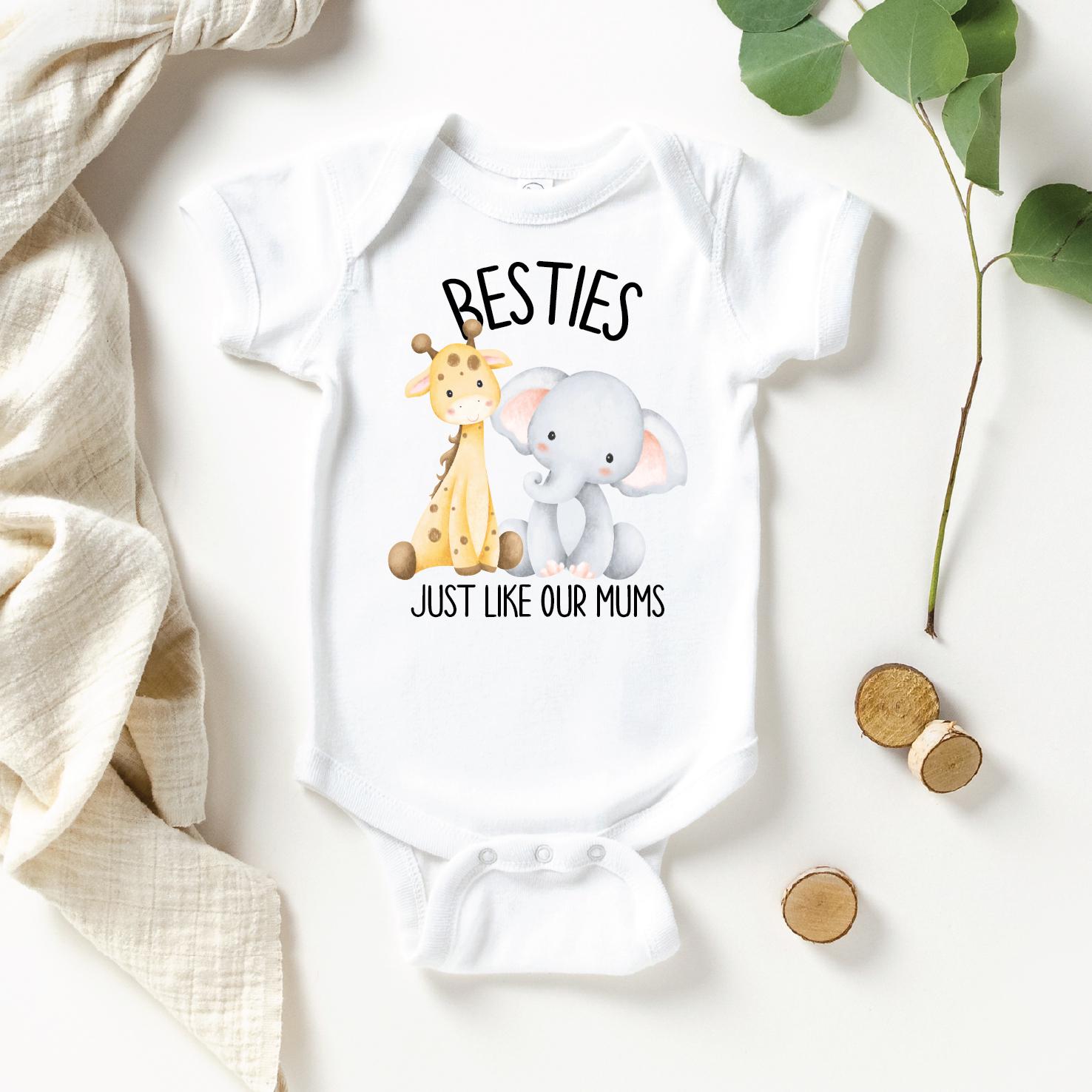 Two young girls sitting on the grass, wearing matching white t-shirts with a giraffe and elephant design and the text "Besties Just like our Mums".