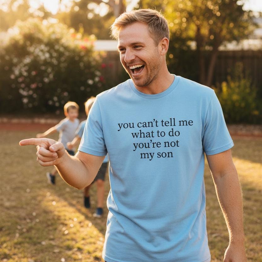 Laughing dad in a backyard wearing a light blue t-shirt with black serif text reading You Can't Tell Me What To Do You're Not My Son.