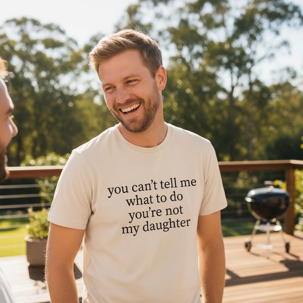 Laughing dad on an outdoor deck wearing a natural cream t-shirt with black serif text reading You Can&amp;#39;t Tell Me What To Do You&amp;#39;re Not My Daughter.
