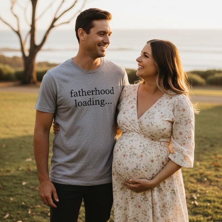 Smiling expecting couple standing together at sunset by the coast, dad-to-be wearing a grey marle t-shirt with black lowercase serif text reading fatherhood loading.