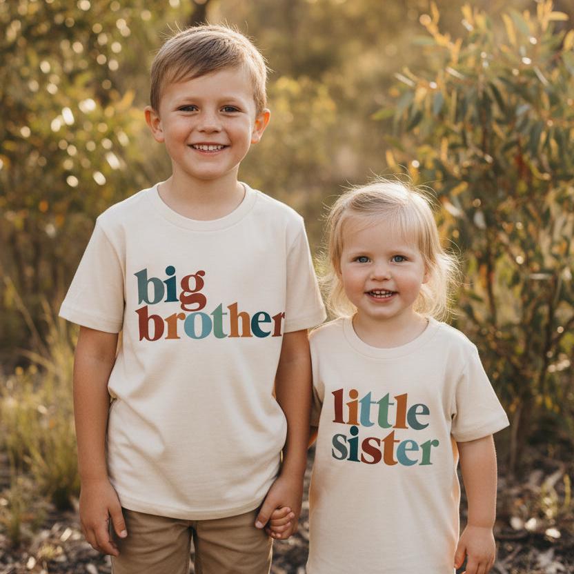 Siblings wearing matching natural cream t-shirts with retro multi-colour lettering reading Big Sister, Little Brother, Big Brother and Little Sister, photographed outdoors in golden afternoon light.