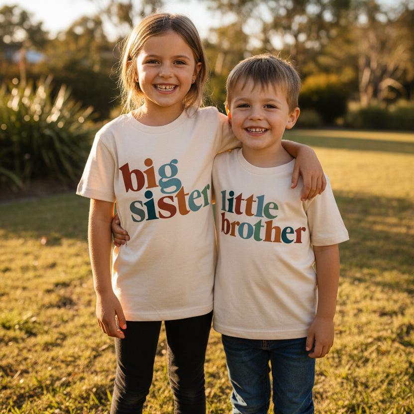 Siblings wearing matching natural cream t-shirts with retro multi-colour lettering reading Big Sister, Little Brother, Big Brother and Little Sister, photographed outdoors in golden afternoon light.