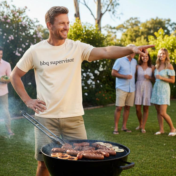 Smiling man pointing while standing at a barbecue in a backyard, wearing a natural cream t-shirt with black lowercase serif text reading bbq supervisor.