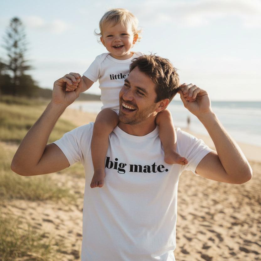 Tilly&amp;Wilbur white matching set — an adult tee printed with &amp;quot;big mate.&amp;quot; and a baby bodysuit printed with &amp;quot;little mate.&amp;quot; in minimalist black lowercase text, worn by a dad carrying his baby on the beach.