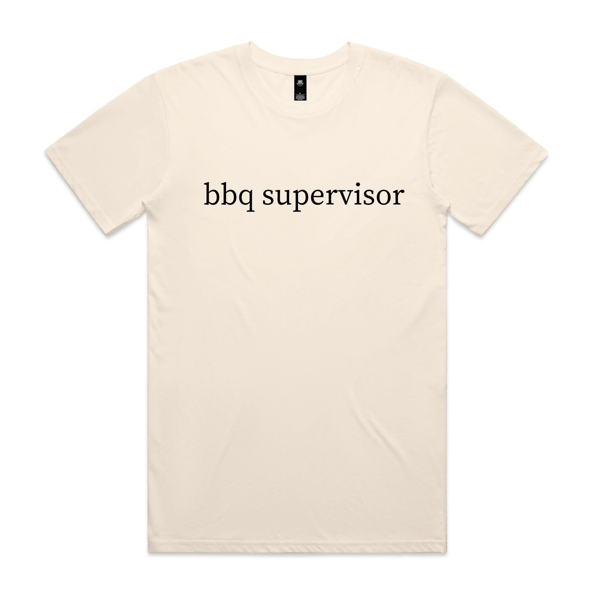 Smiling man pointing while standing at a barbecue in a backyard, wearing a natural cream t-shirt with black lowercase serif text reading bbq supervisor.