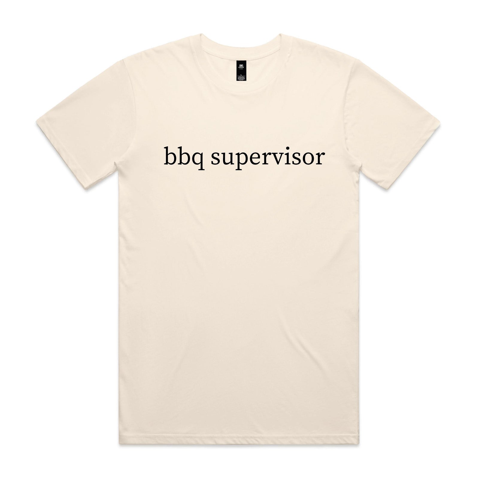 Smiling man pointing while standing at a barbecue in a backyard, wearing a natural cream t-shirt with black lowercase serif text reading bbq supervisor.