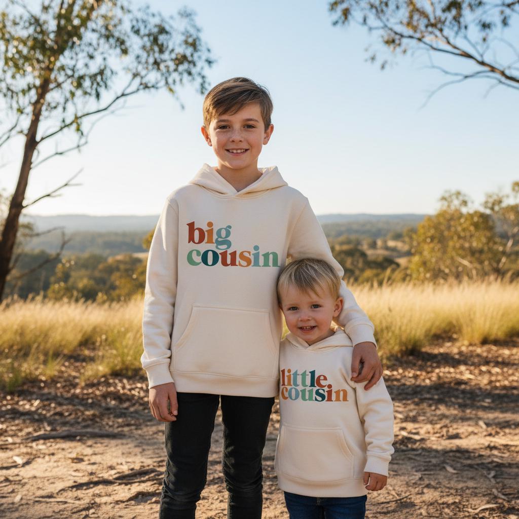 Two young cousins standing in the Australian bush wearing cream Big Cousin and Little Cousin hoodies with colourful retro lettering