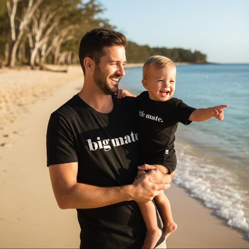 Tilly&Wilbur black matching set — an adult tee printed with "big mate." and a baby bodysuit printed with "little mate." in minimalist white lowercase text, worn by a dad carrying his baby on the beach.