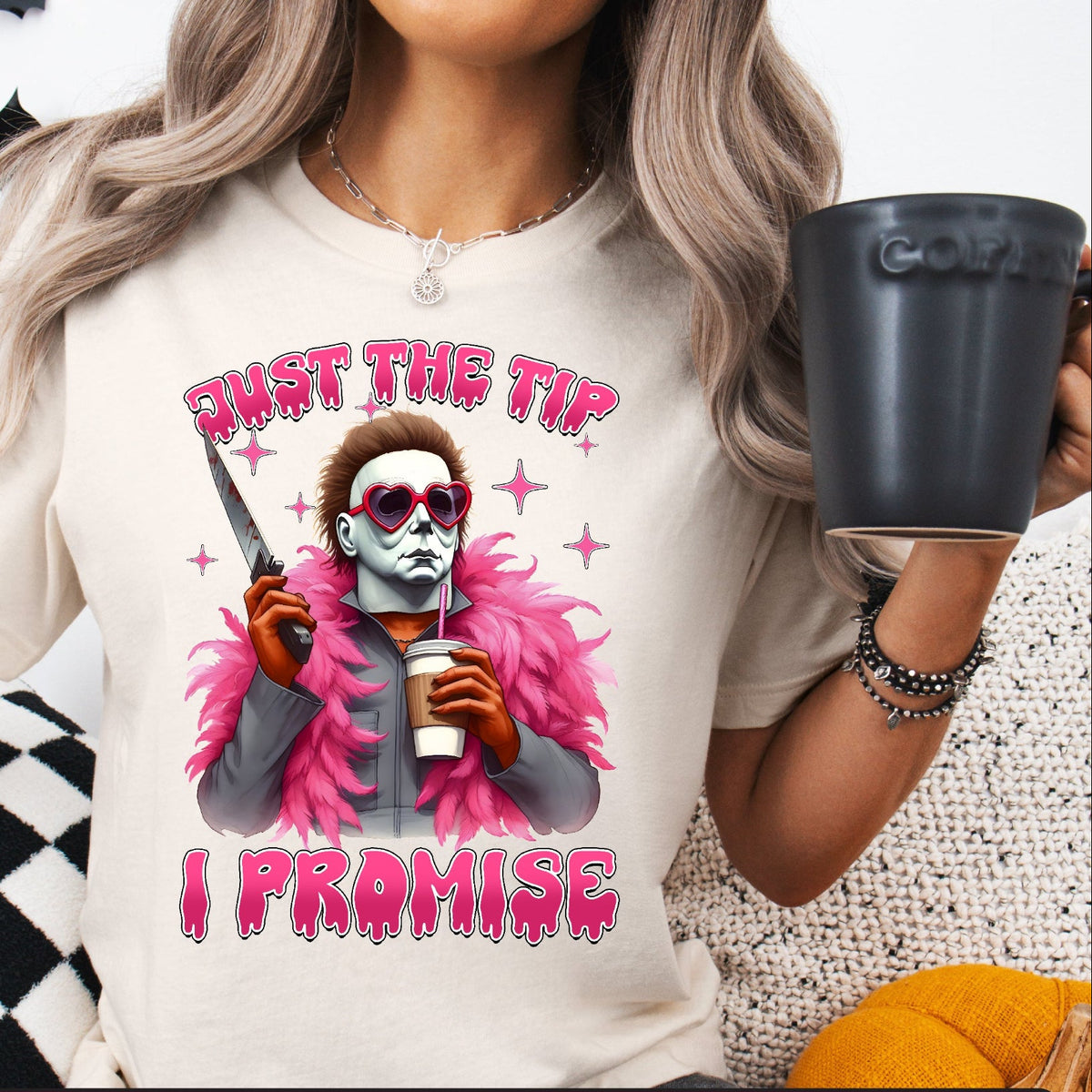 A person wearing a white T-shirt with a graphic print of Michael Myers from Halloween, holding a coffee cup, and text &#39;JUST THE TIP, I PROMISE&#39;.