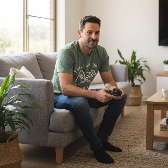 A sage green short-sleeve t-shirt with a white graphic print on the front featuring a video game controller and the text 'LEVELLED UP TO DADDY'.