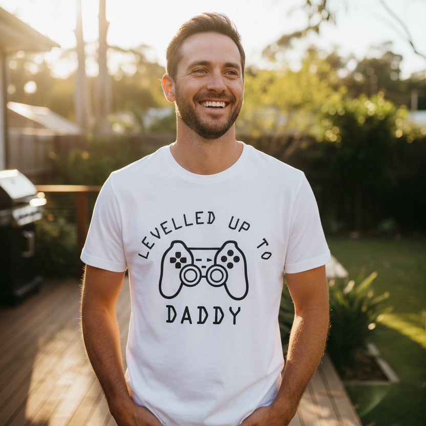 A white short-sleeve crewneck t-shirt with a black graphic print on the front. The graphic features a video game controller flanked by the text 'LEVELLED UP TO' above and 'DADDY' below, set against a plain white background.