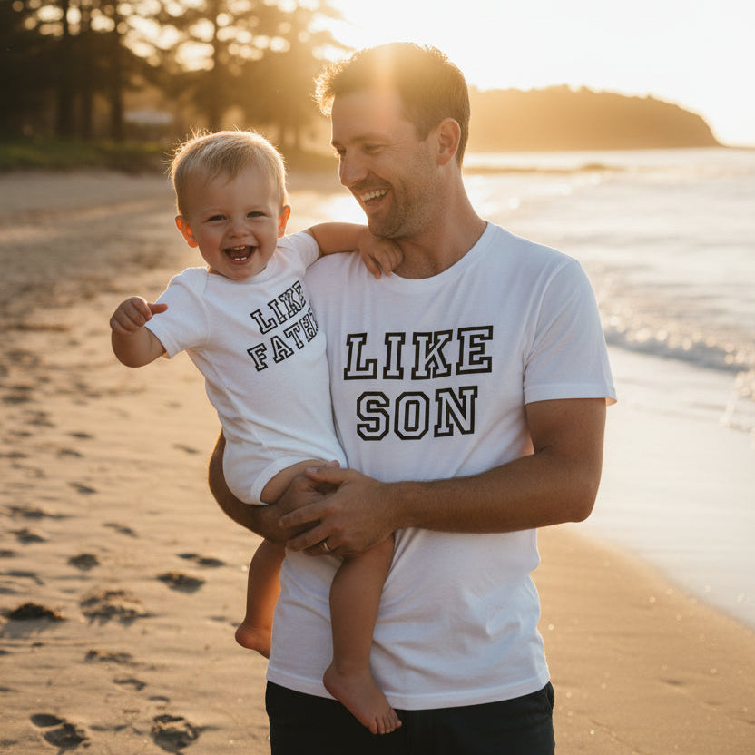 A white short-sleeve t-shirt and a matching white short-sleeve baby bodysuit displayed on a plain white background. The t-shirt features the text 'LIKE SON' in black varsity-style lettering, and the bodysuit features 'LIKE FATHER' in the same black lettering.