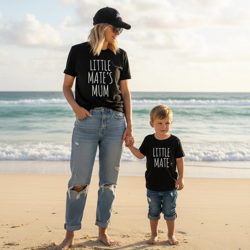 Mother and child wearing matching black Little Mate’s Mum and Little Mate shirts, walking together on the beach