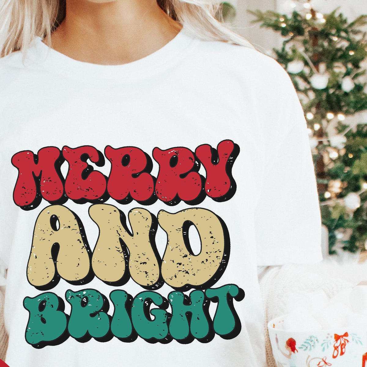A person holding a mug and wearing a white crew neck t-shirt with the text 'Merry and Bright' in multicolored letters.