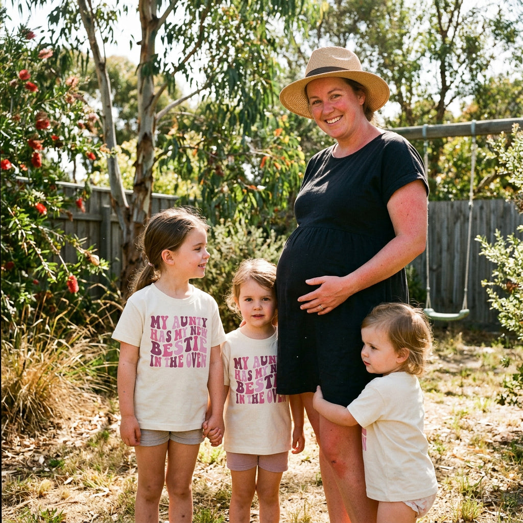 A cream-colored short-sleeve t-shirt with a pink graphic text that reads 'MY AUNTY HAS MY NEW BESTIE IN THE OVEN'. 