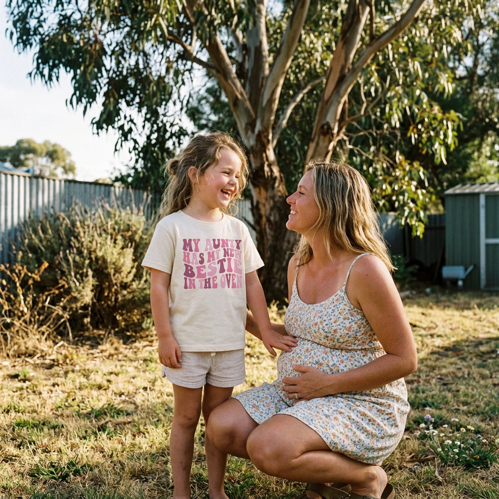 A cream-colored short-sleeve t-shirt with a pink retro-style text graphic reading 'MY AUNTY HAS MY NEW BESTIE IN THE OVEN'.