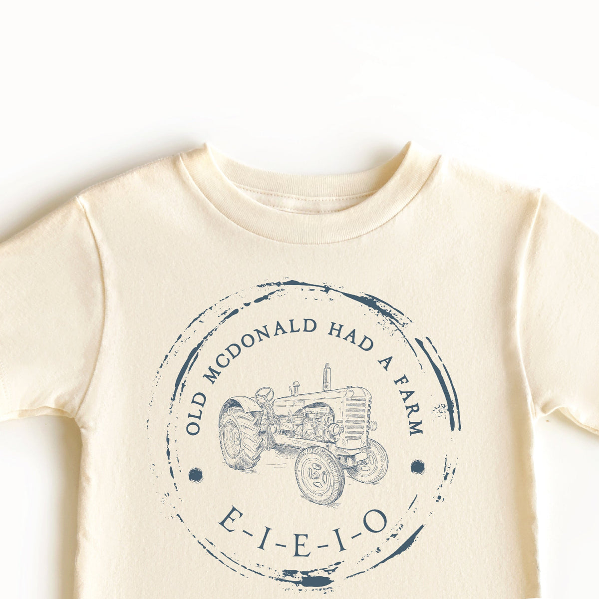 A beige-colored toddler t-shirt with a graphic print of &#39;Old McDonald Had a Farm&#39; including a tractor illustration and the text &#39;E-I-E-I-O&#39;.