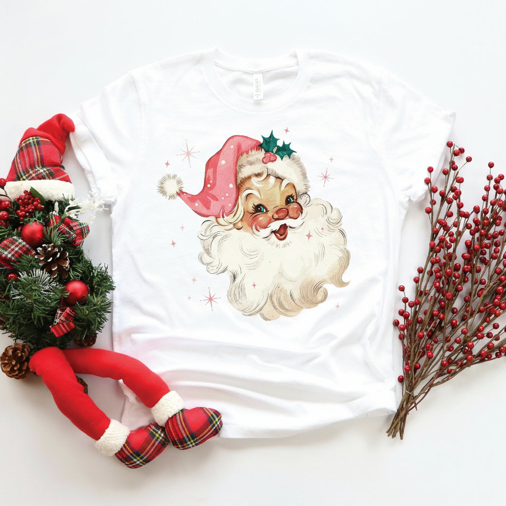 A graphic t-shirt with a vintage Santa Claus design, featuring a pink hat, displayed with Christmas decorations and text indicating it is suitable for babies, kids, and adults.