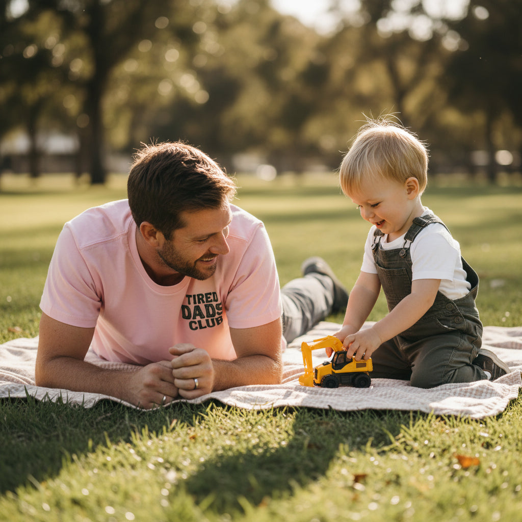 A light pink short-sleeve t-shirt shown from both the front and back. The front features the text 'TIRED DADS CLUB' in dark green on the left chest.