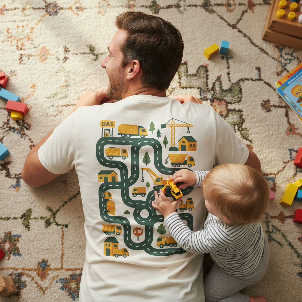 An off-white short-sleeve t-shirt with a large graphic print on the front depicting a winding road filled with construction vehicles, trucks, cranes, and trees in yellow, green, and dark gray.