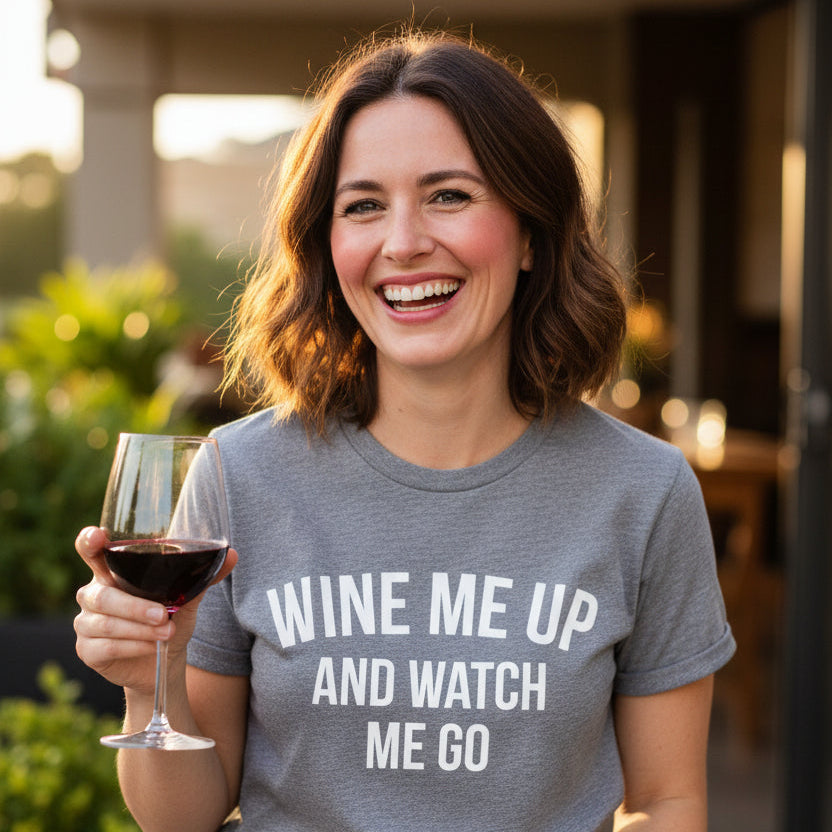 Smiling woman holding a glass of red wine outdoors wearing a white Wine Me Up And Watch Me Go t-shirt with bold black lettering