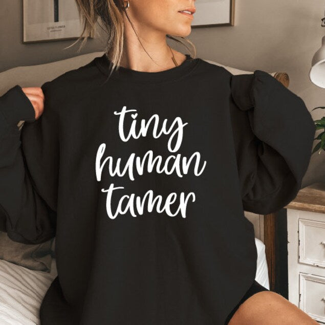 A person wearing a black sweatshirt with the text &#39;TINY HUMAN TAMER&#39; printed on the front.